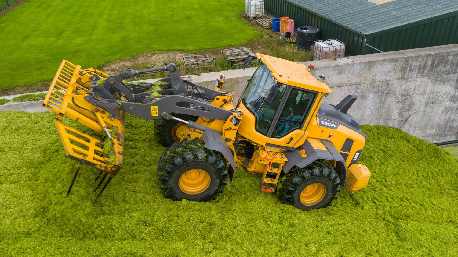 Heavy duty construction loader with yellow fork attachment lifting equipment on grassy terrain next to buildings.
