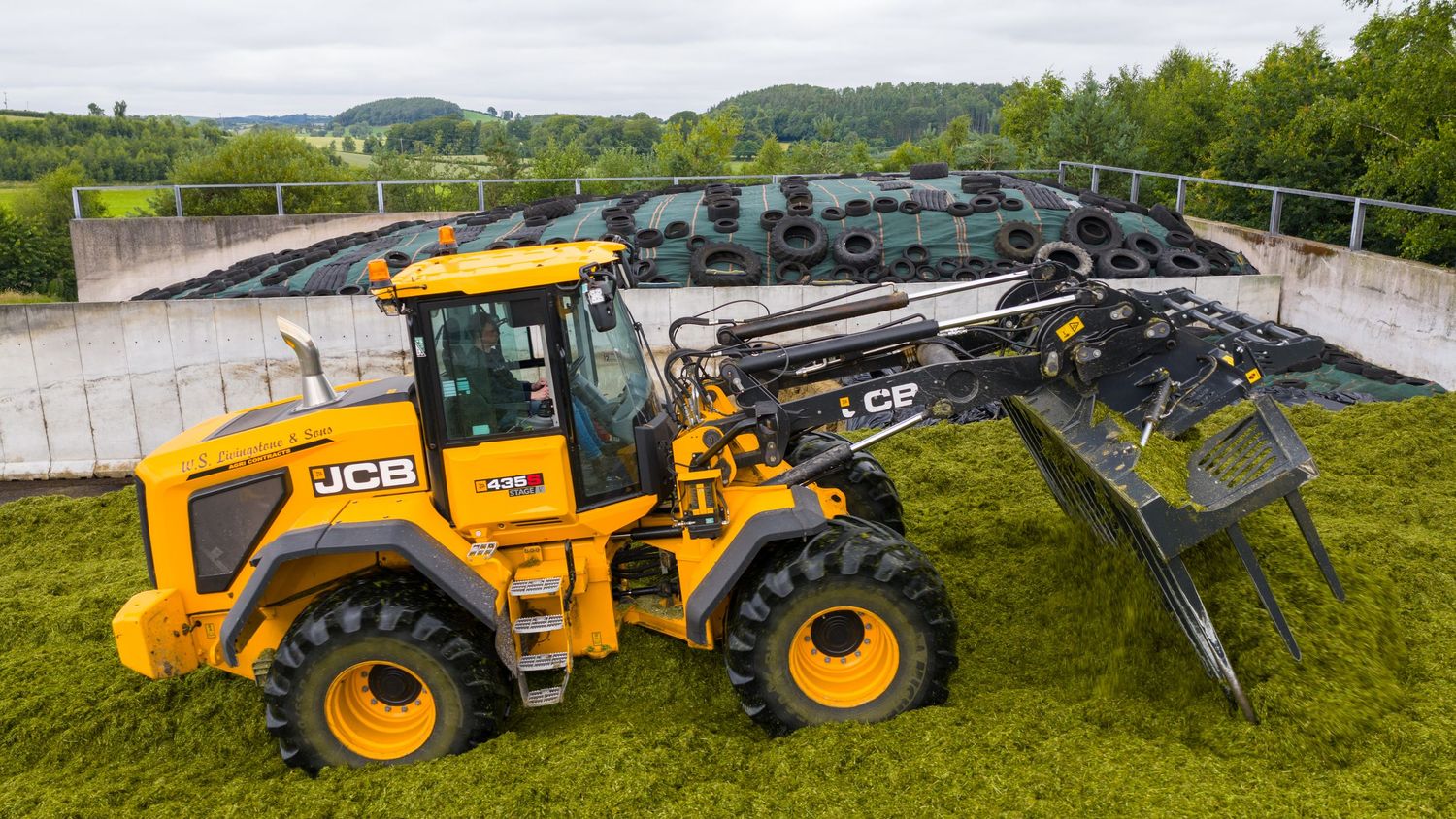 Row of heavy construction equipment including JCB rollers and tractors parked on grass against rural landscape backdrop.