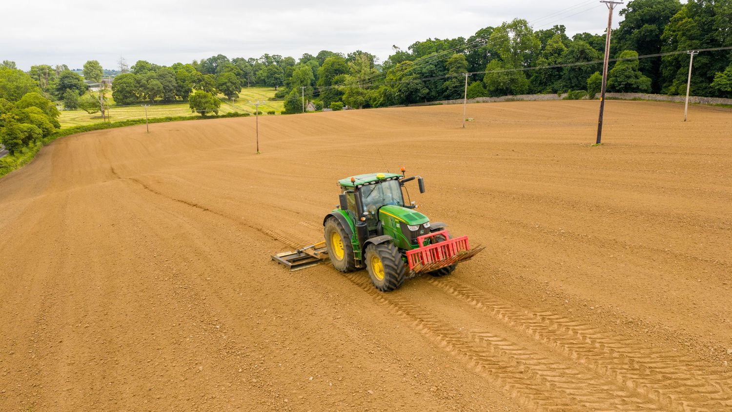 Aerial view of green tractor tilling dry brown soil in agricultural field with power lines above.