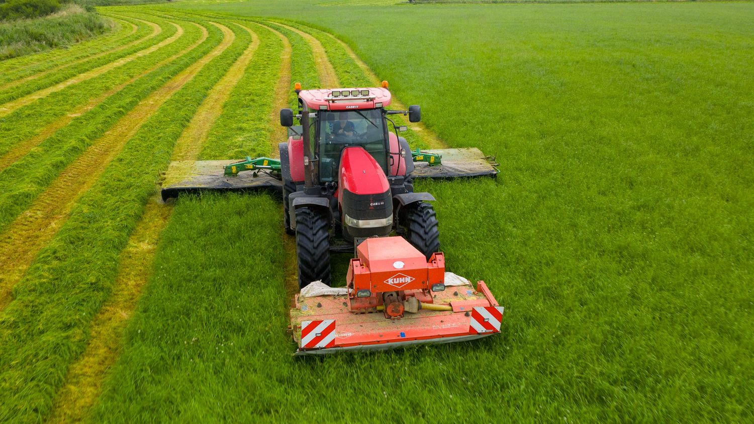 A red tractor mowing green grass in neat rows with a large mowing attachment creating parallel stripes in a field.