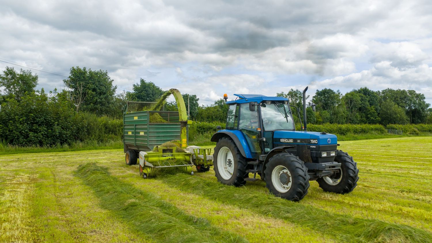 Blue tractor pulling green hay baler across sunny grass field under cloudy summer sky.