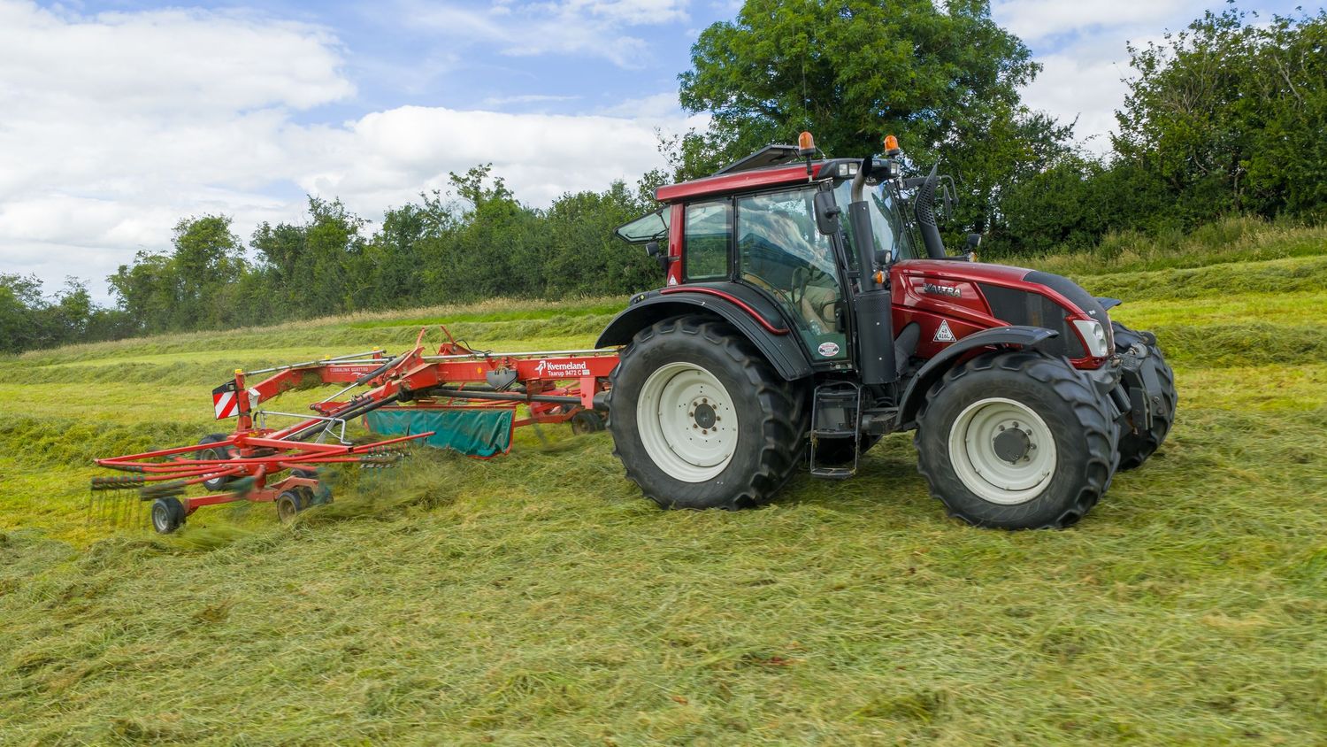 Modern agricultural tractors working in a lush green field pulling hay raking equipment on a sunny day.