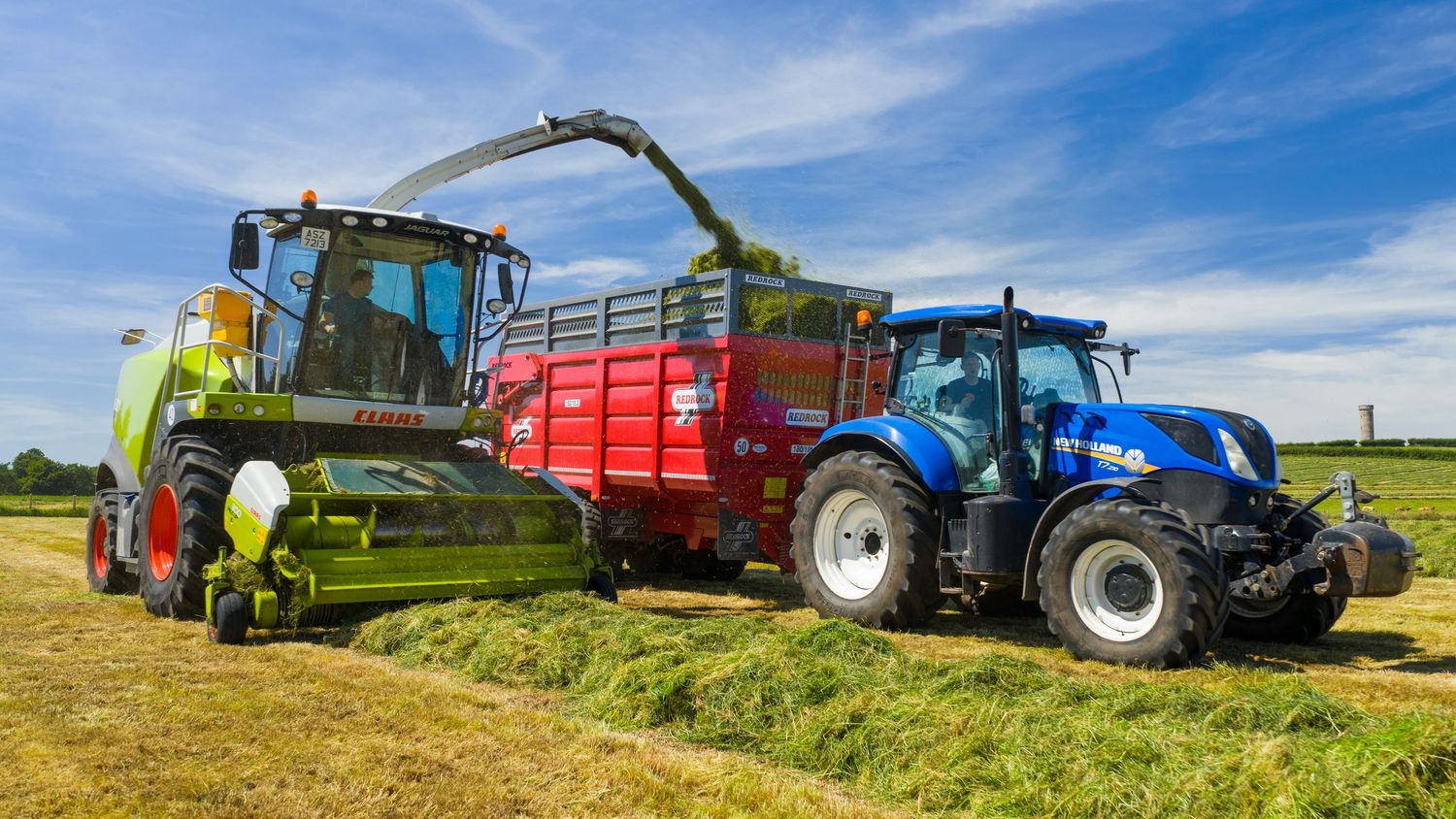 Modern farming machinery collecting and loading freshly cut silage into a red trailer during harvest season.