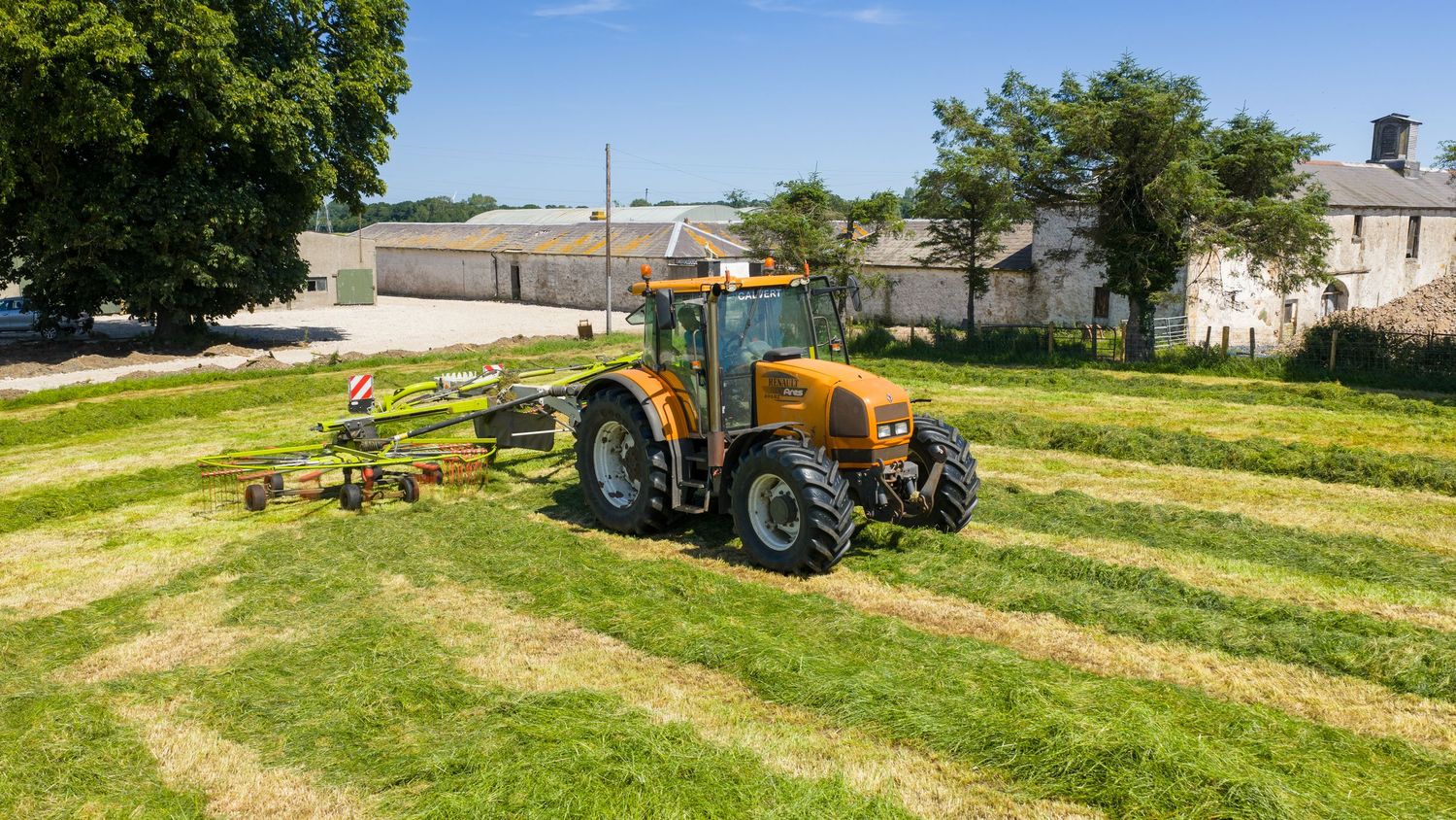 Yellow tractor cutting grass in parallel rows on a sunny day at a countryside property.