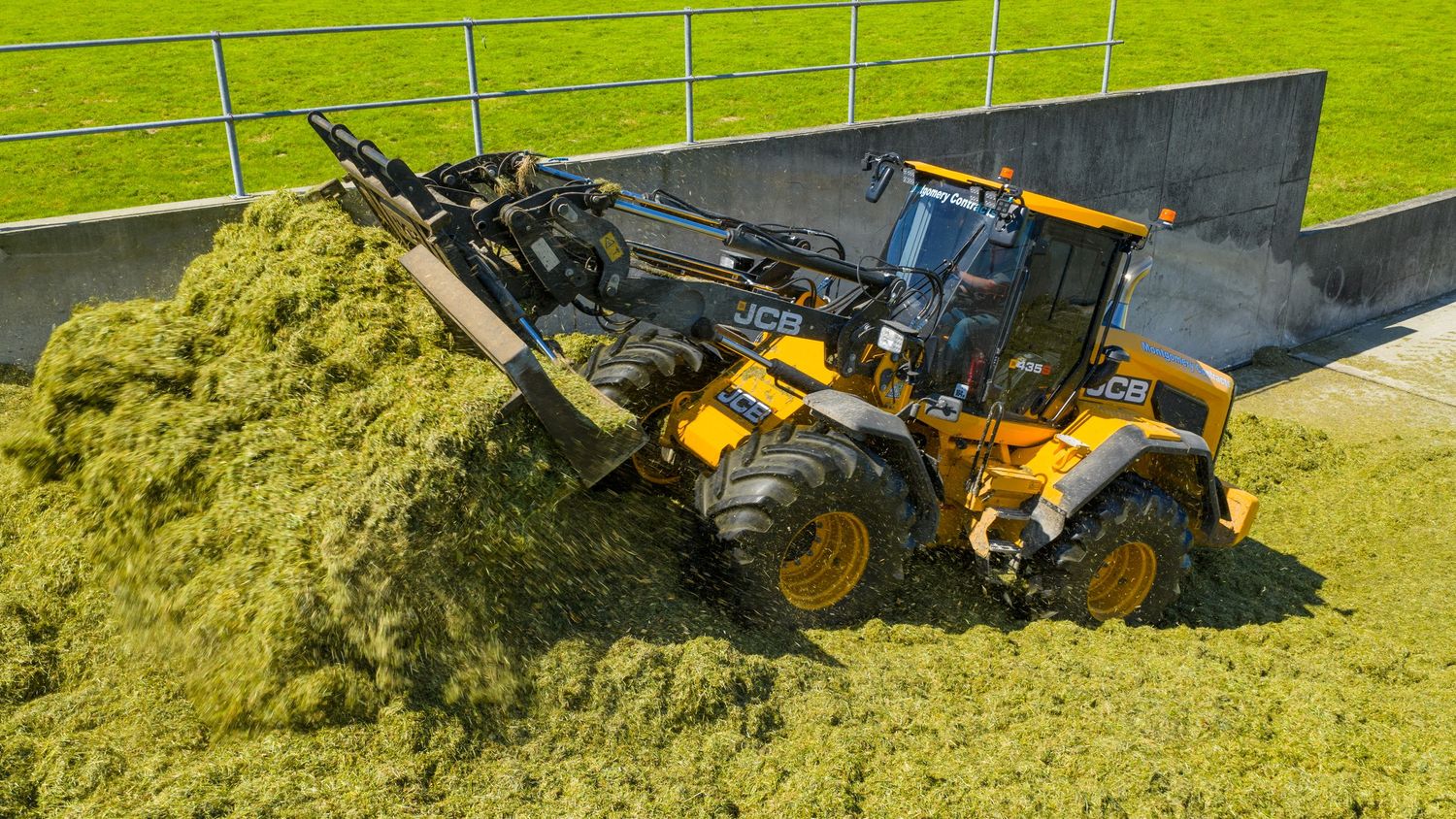 Yellow construction loader pushes large pile of green silage on dairy farm with grassy field in background.