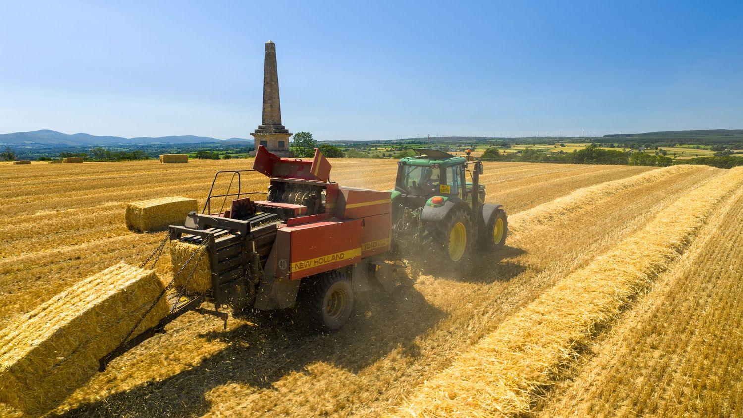 Tractor and hay baler working together in golden wheat field during summer harvest under blue sky.