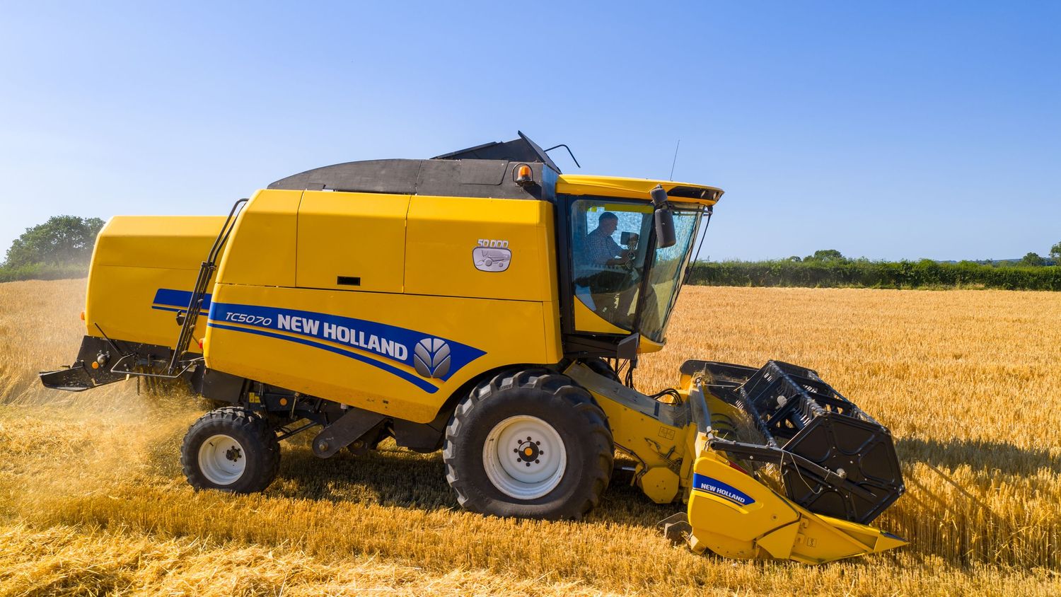 Yellow New Holland combine harvester working in golden wheat field during summer harvest season.