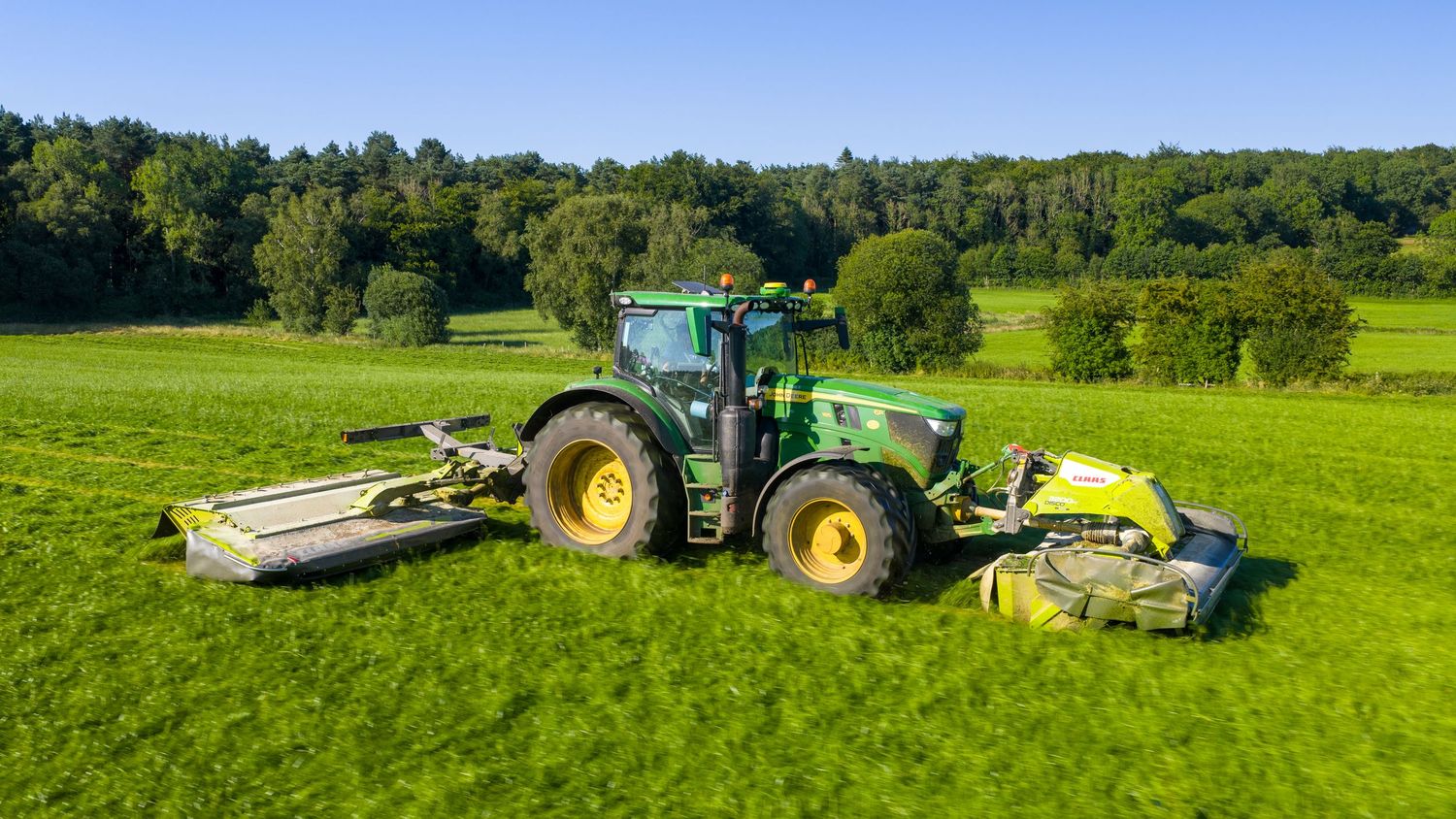 A green John Deere tractor with agricultural equipment moves across vast rolling farmland under a bright blue sky.