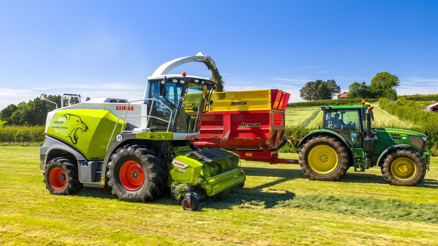 Large green forage harvester working alongside a red trailer and green tractor in a sunny agricultural field.