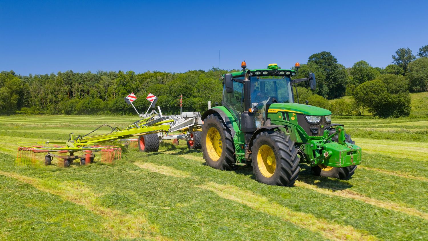 Green John Deere tractor raking fresh cut hay in a lush summer field on a sunny day.