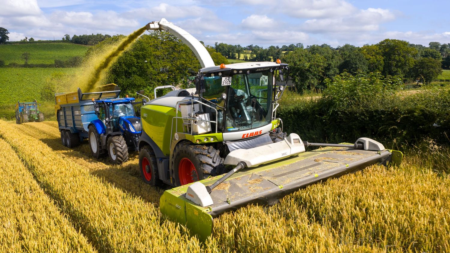 Blue tractor with disc mower cutting grass on a sloped agricultural field on a sunny day with puffy clouds.