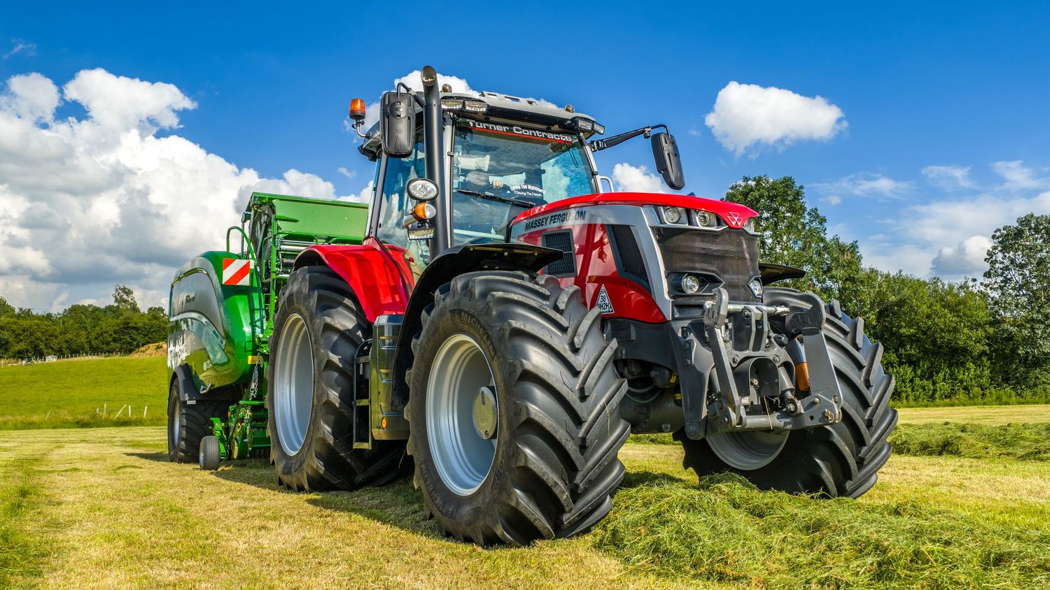 Modern red and black agricultural tractor with trailer working in sunny green field under blue sky with white clouds.