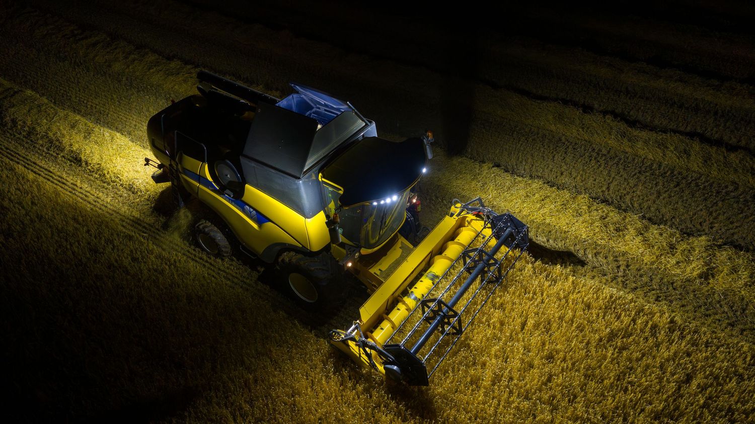 Aerial night view of a combine harvester with bright yellow lights illuminating golden crops during harvest.