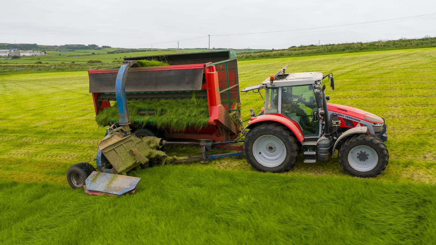 Red tractor pulling harvesting equipment across lush green farmland on an overcast day.