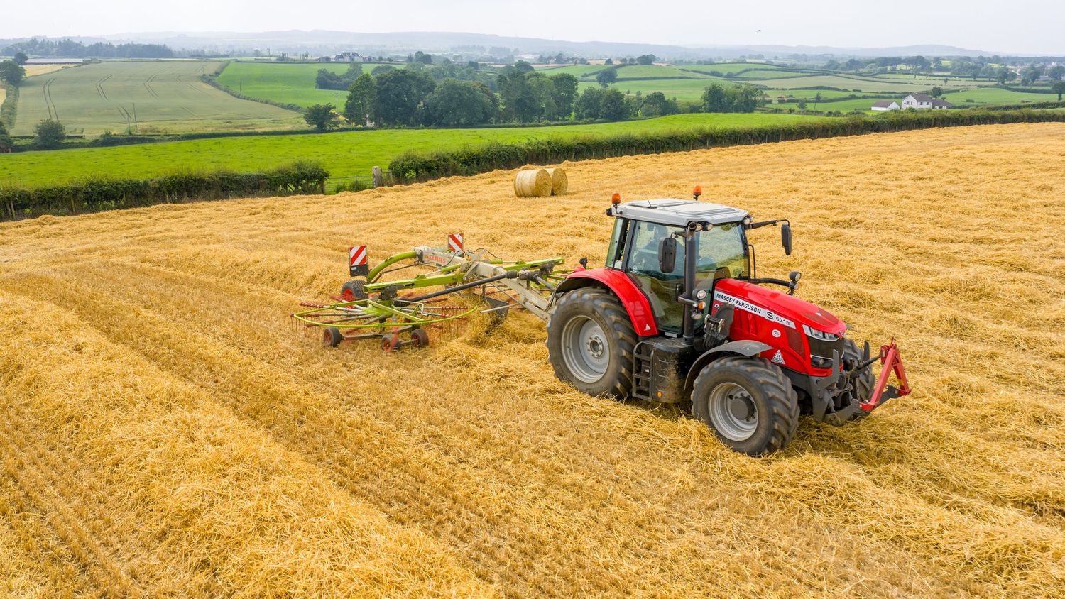 Red tractor pulling agricultural equipment across golden harvested wheat field on sunny summer day.