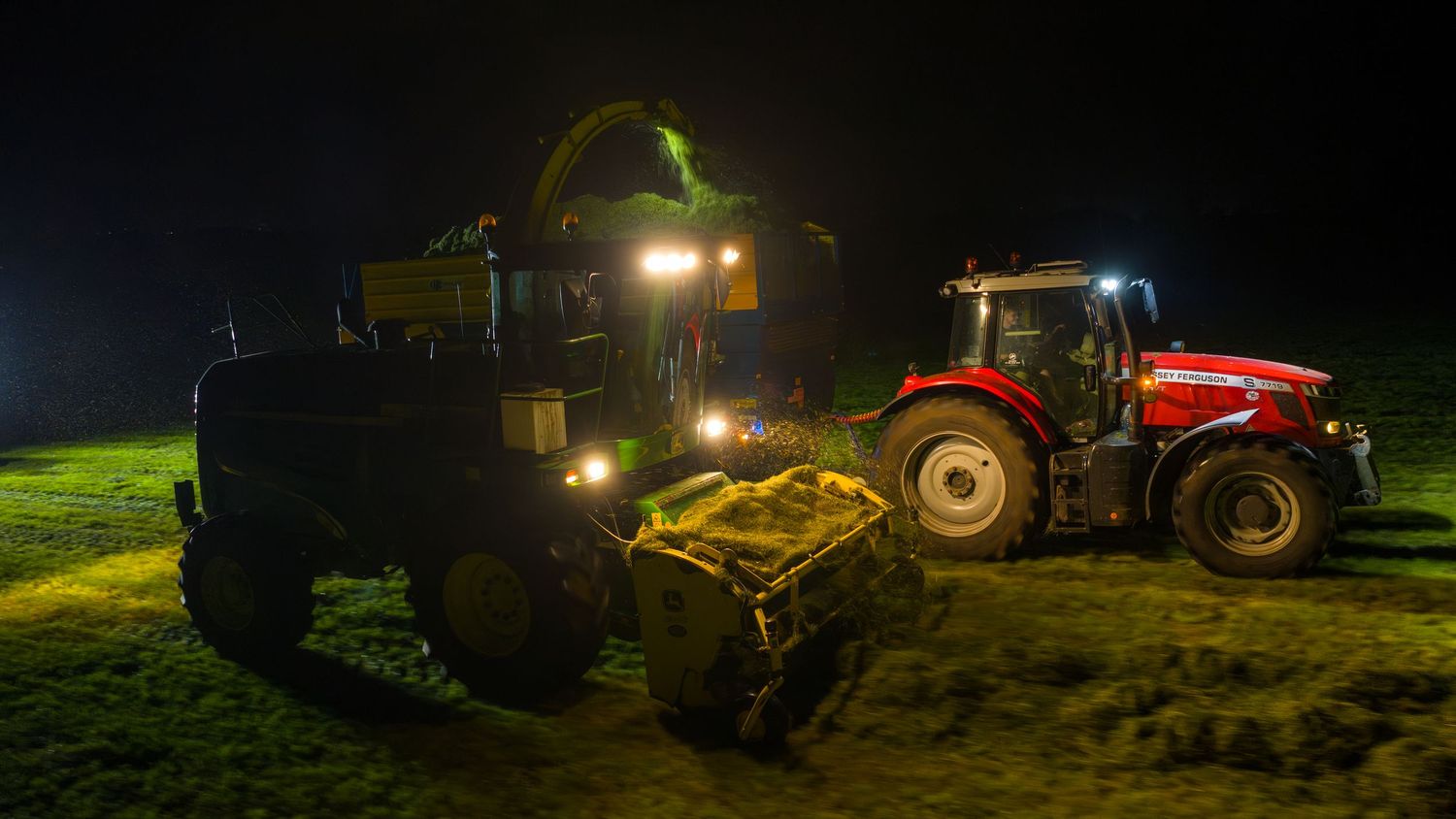 Agricultural tractors and farm machinery operating at night with bright headlights illuminating dark field.