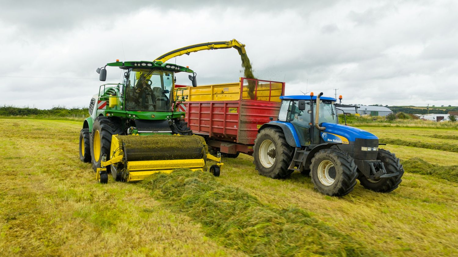 A green harvester loads silage into a red trailer pulled by a blue tractor in a grassy field under cloudy skies.