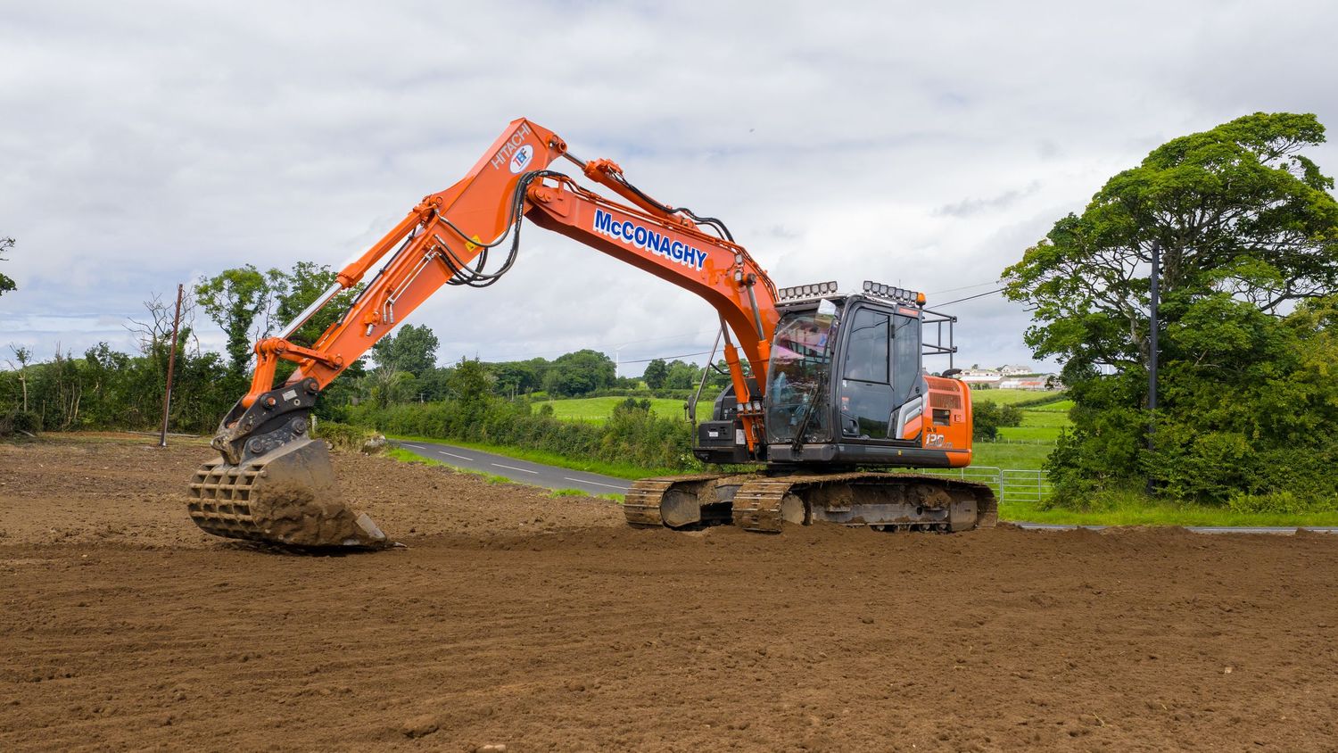 Large orange excavator with bucket arm extended on cleared brown dirt field against green trees and cloudy sky.