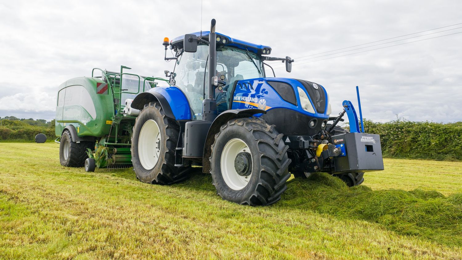 Blue New Holland tractor pulling green fertilizer spreader on grassy field under cloudy sky.
