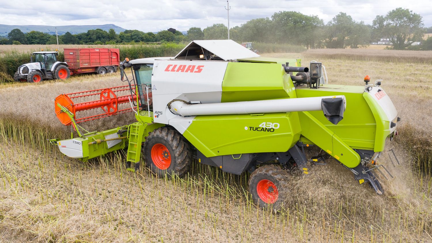 Green and white Claas combine harvester working in a grain field with red trailer behind.