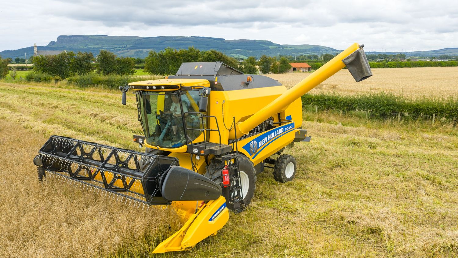 Modern yellow combine harvester working in a rural wheat field with mountains visible in the background.