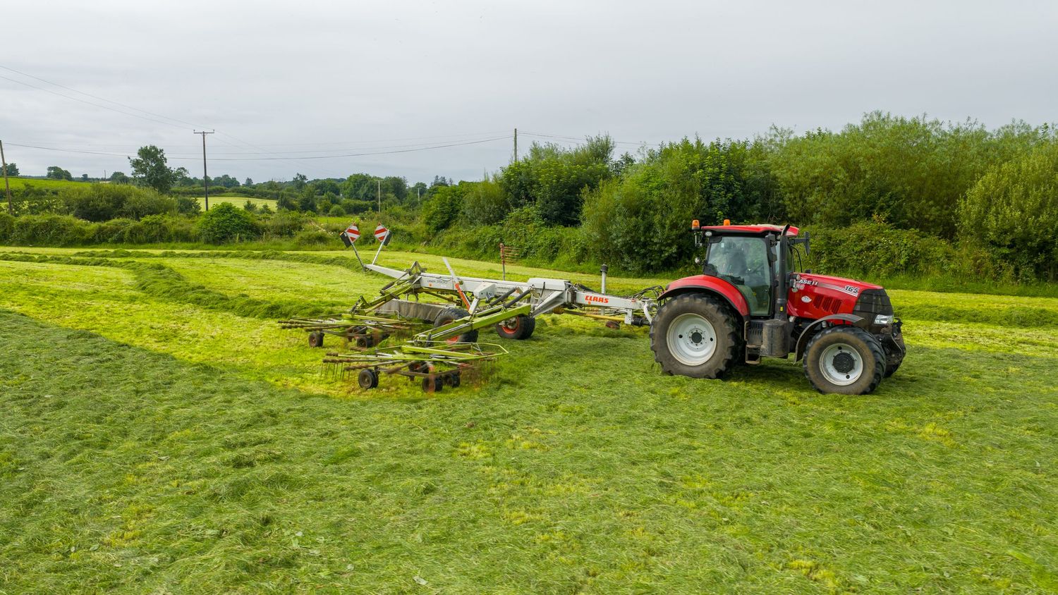 Red tractor pulling farm equipment through a lush green field with yellow wildflowers on an overcast day.