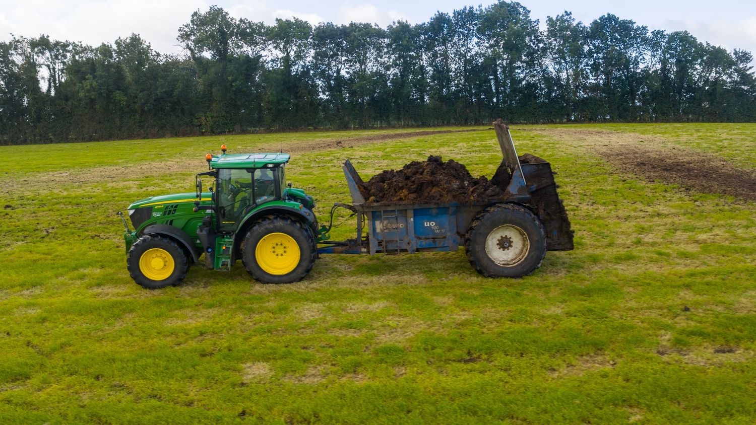 Green John Deere tractor with manure spreader working in a grassy field with treeline in background.