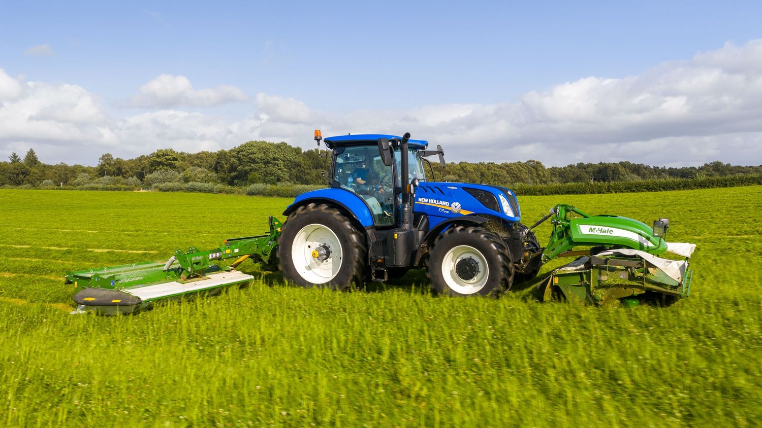 A panoramic view shows blue tractors working in green agricultural fields under a partly cloudy sky.
