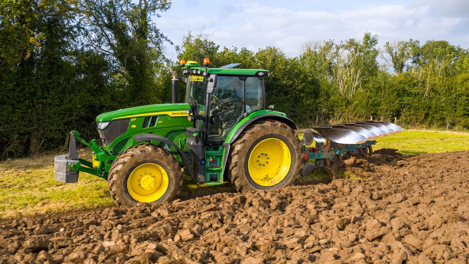 Modern green John Deere tractor plowing agricultural field with brown soil and trees in background.