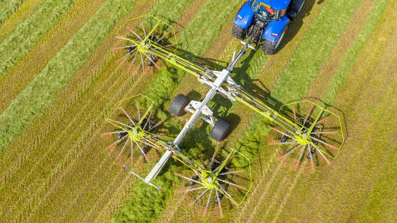 Aerial view of a blue tractor with spinning hay rake machinery working in a green agricultural field.