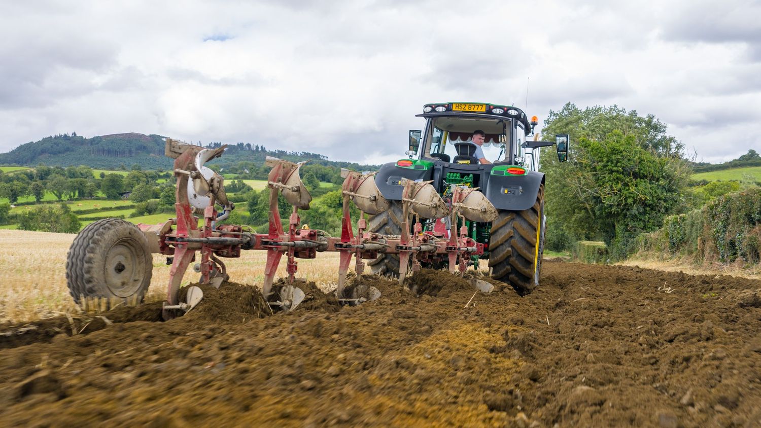 Blue tractor pulling a large plow through brown soil in a rural agricultural field with green hills in the background.