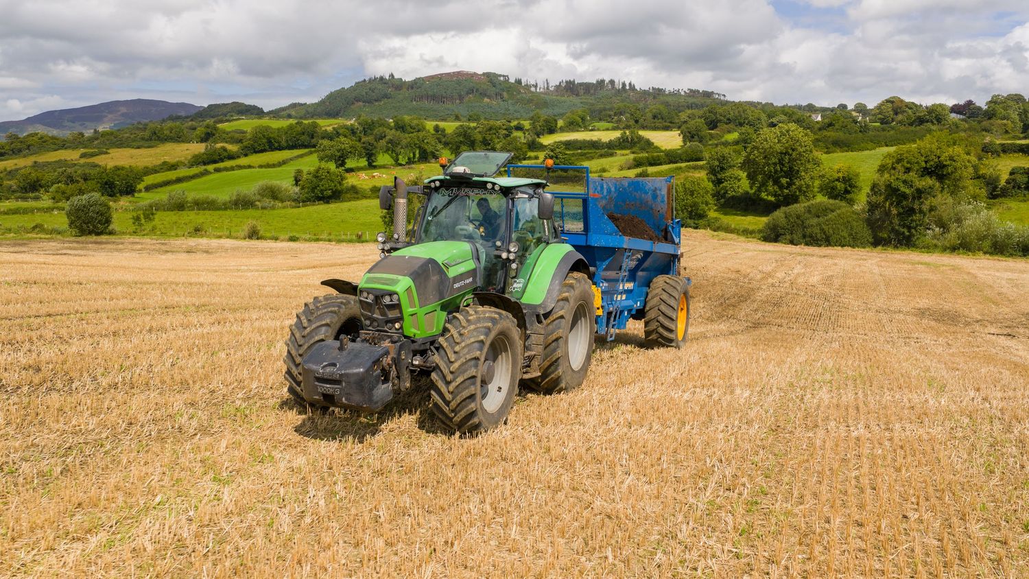 A line of modern tractors working in farming fields with rolling green hills and cloudy skies in the background.
