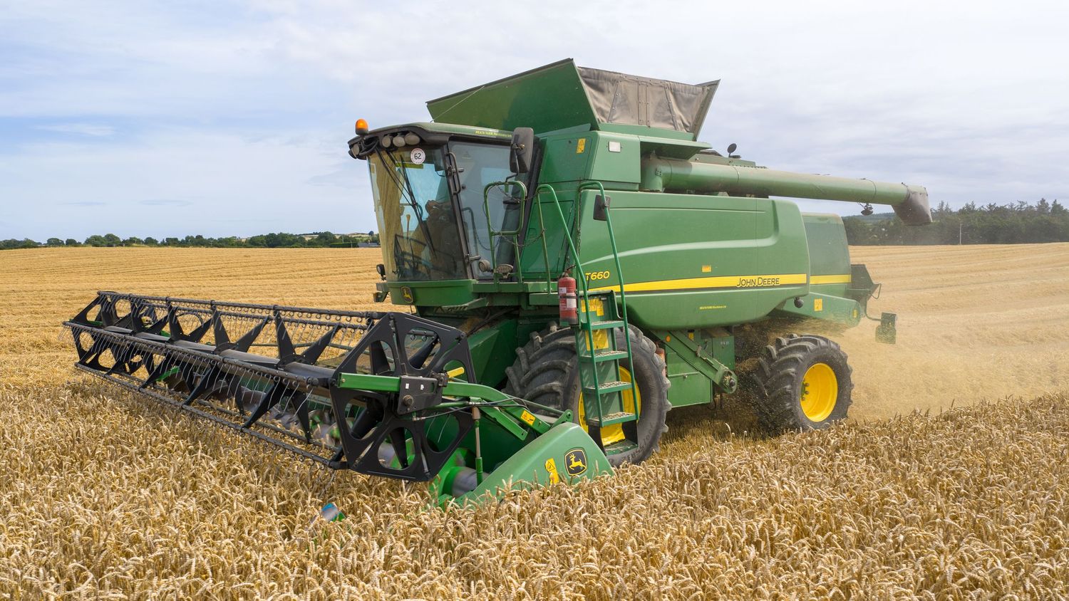 Large green John Deere combine harvester working in golden wheat field during harvest season with extended header.