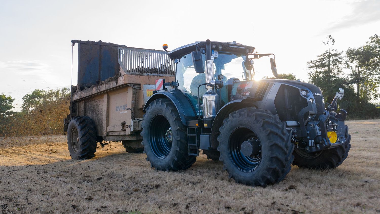 A blue tractor with large wheels parked next to a farm trailer at dusk with its headlights illuminated in a field.