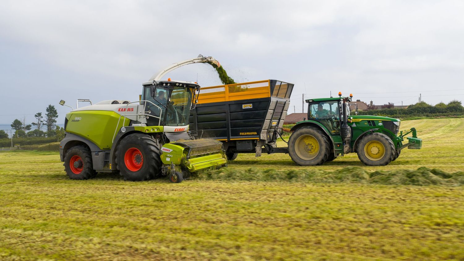 Agricultural machinery including a combine harvester and tractor working together to harvest crops in a green field.
