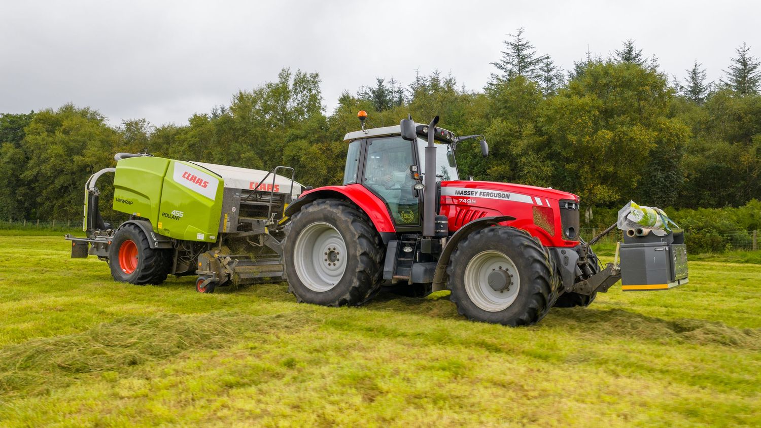 A red Massey Ferguson tractor pulling green farm equipment across a lush grassy field near a treeline.