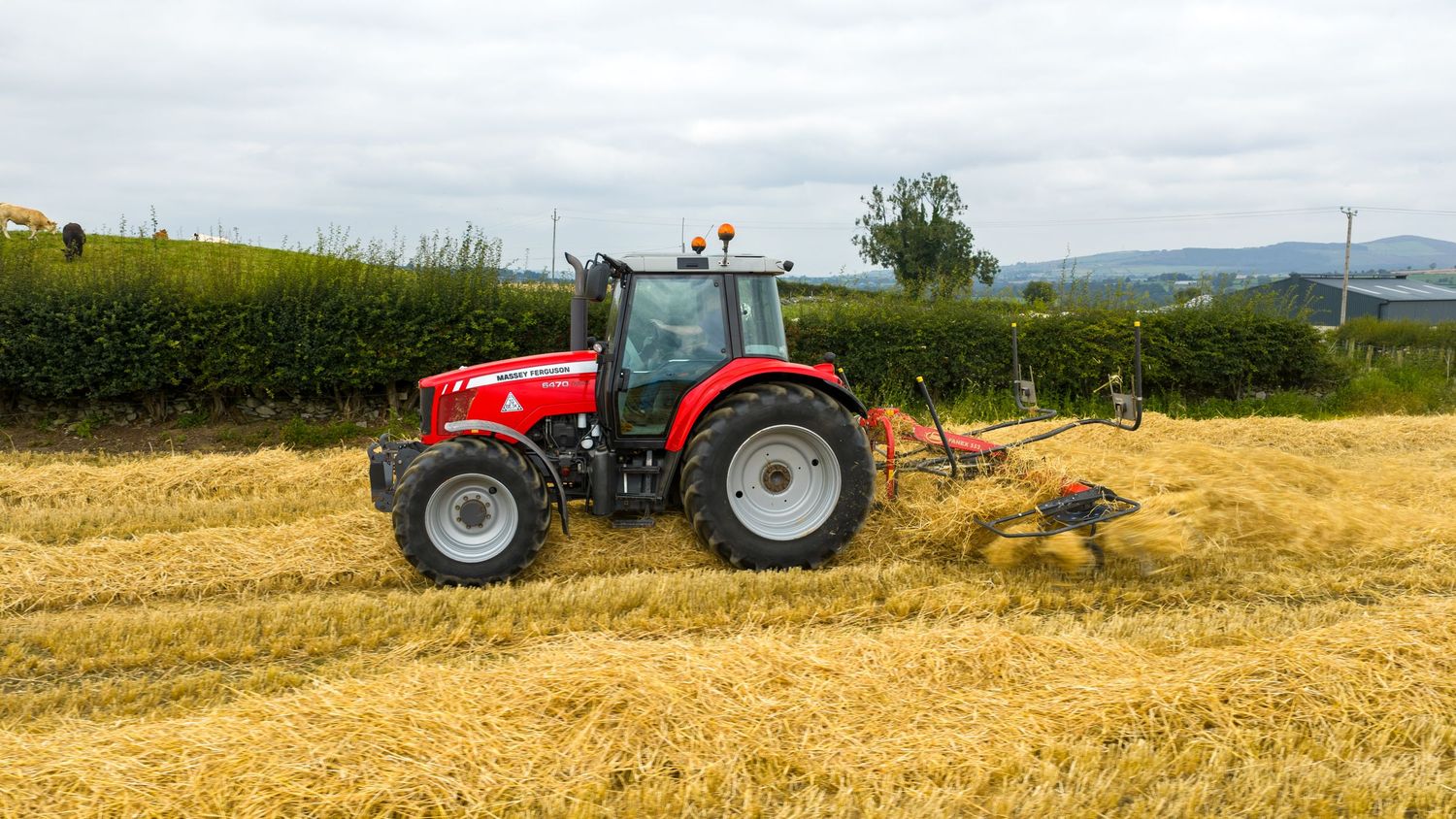 Red Massey Ferguson tractor pulling agricultural equipment through a harvested wheat field on a cloudy day.