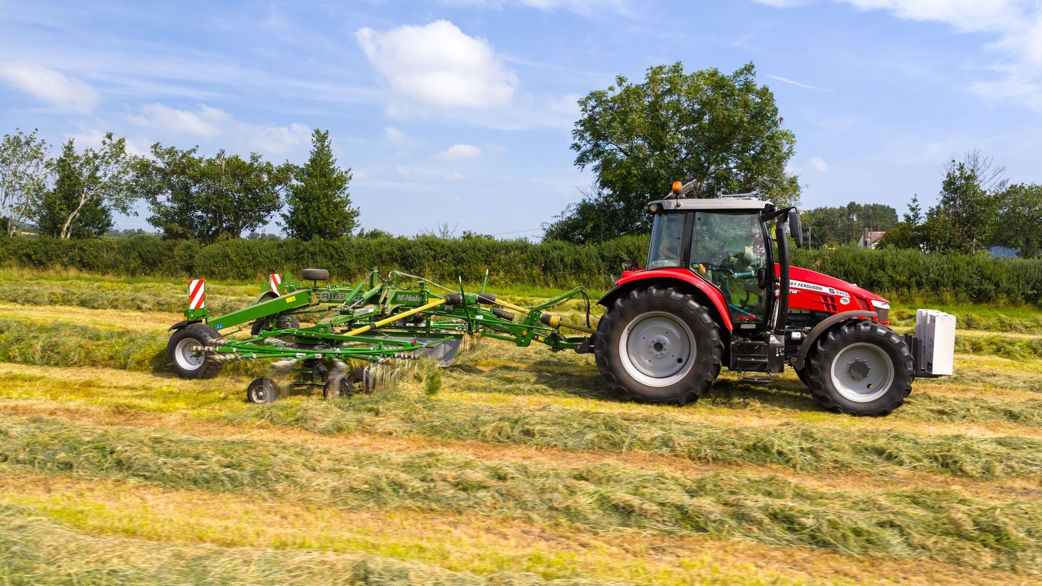 Red tractor pulling green hay raking equipment across a sunny agricultural field.