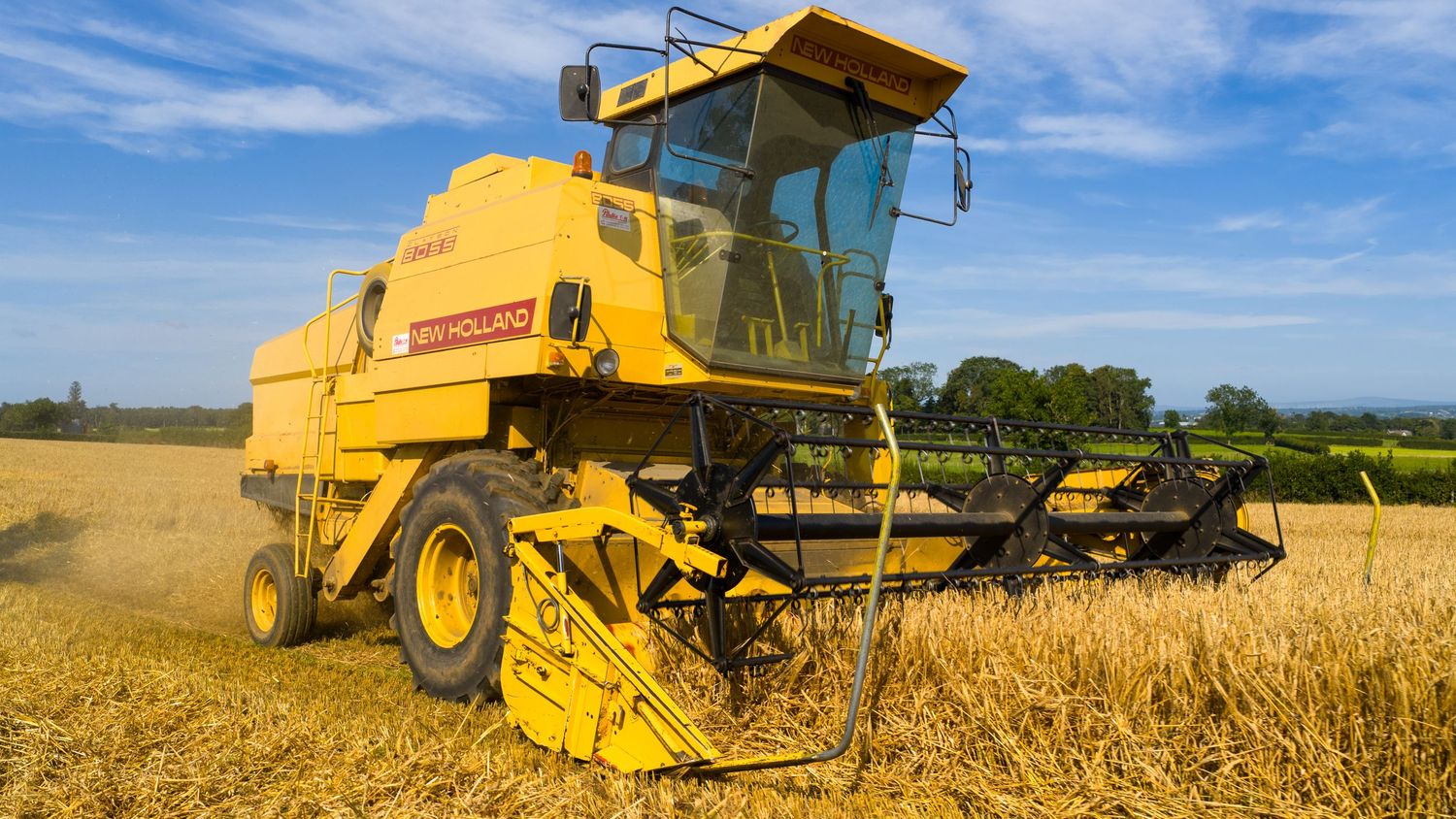 Yellow New Holland combine harvester working in a golden wheat field under blue summer sky.