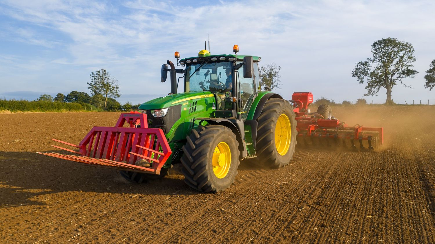 Green John Deere tractor plowing brown field with red attachment under blue sky with scattered clouds.