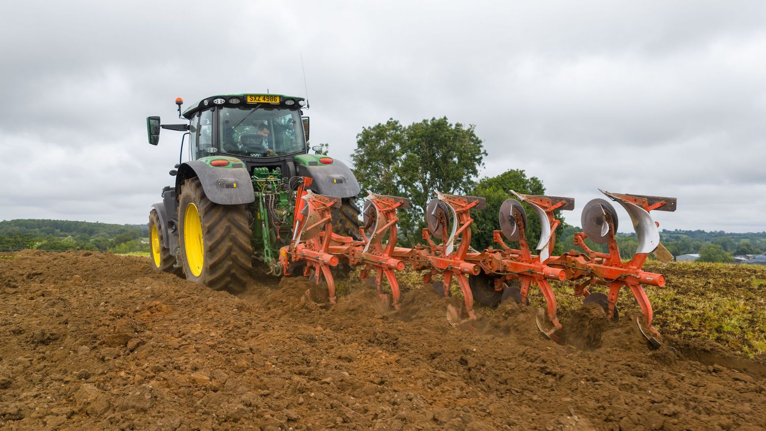 A line of orange agricultural plows move across a farm field under cloudy skies during harvest season.