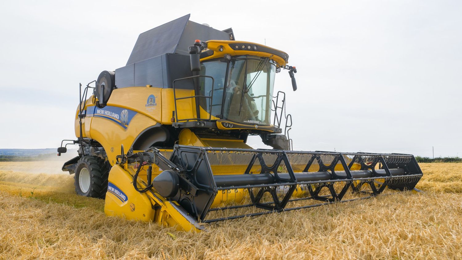Yellow New Holland combine harvester working in golden wheat field with cutting header extended across ripe crop.