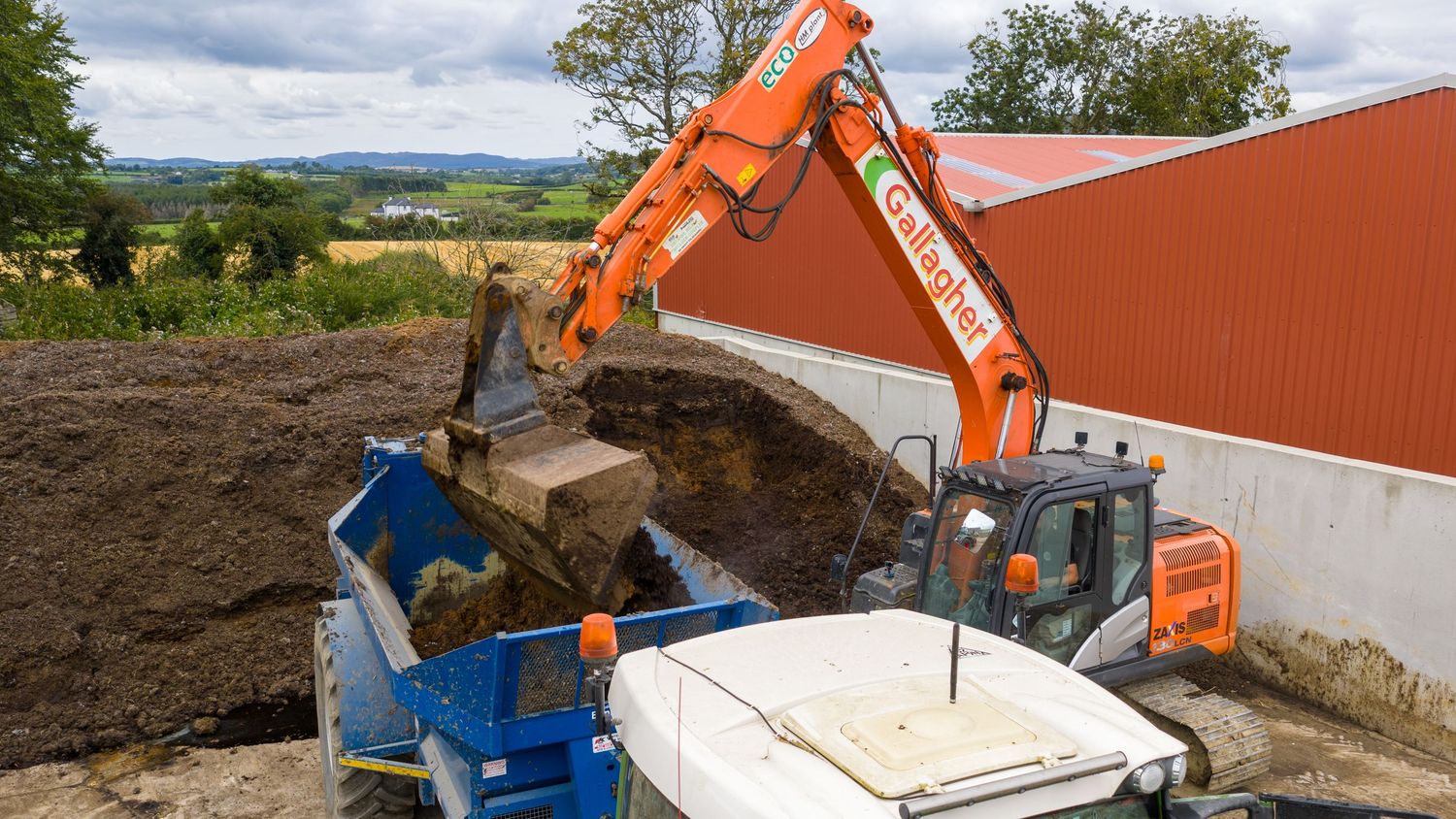 An orange excavator loads dirt into a blue container truck beside a red building.