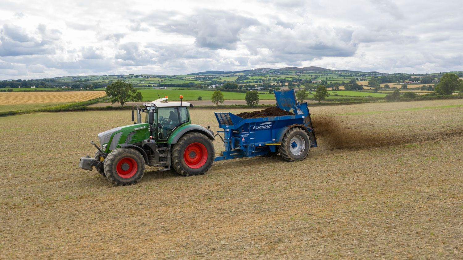 Agricultural tractor and blue machinery working together in a harvested field under cloudy skies.