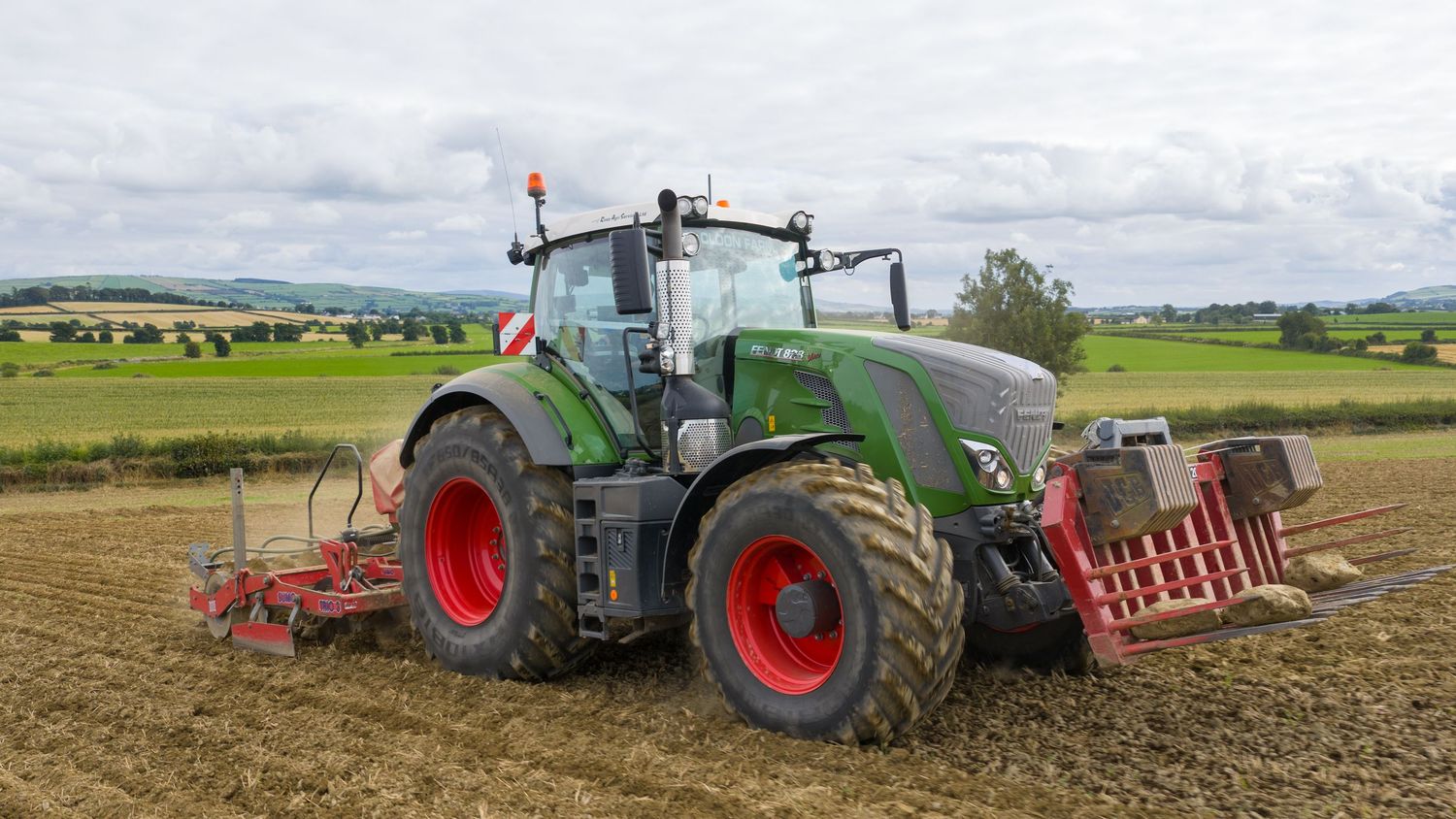 A green Fendt tractor with red wheels plows a field while pulling agricultural equipment on farmland.