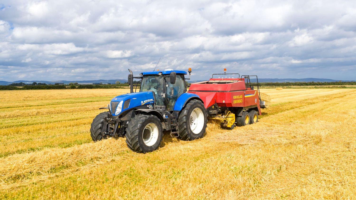 Blue tractor pulling a red baler across golden harvested wheat field under cloudy summer sky.