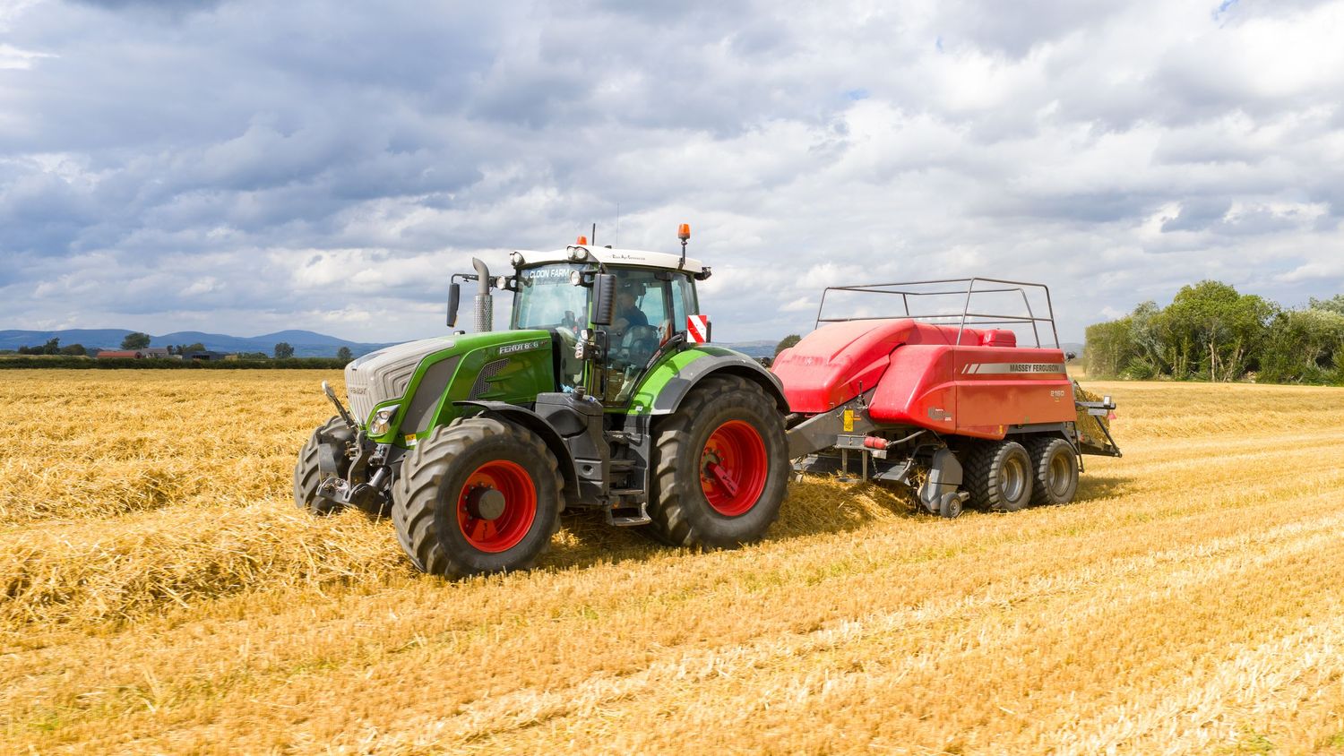 Multiple tractors and combines harvesting golden wheat fields under a cloudy summer sky with rolling hills in background.