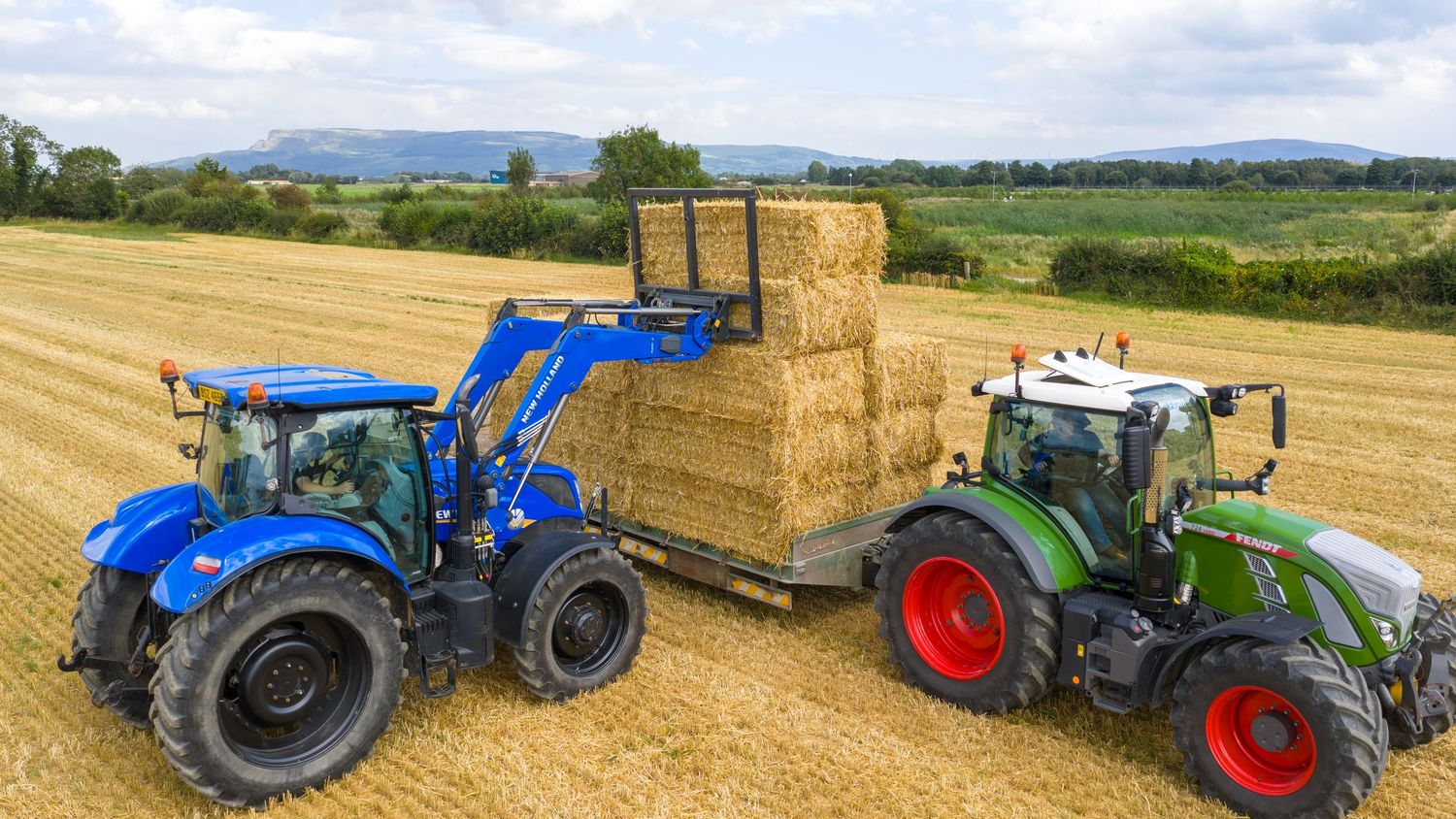 Six agricultural machines and tractors lined up in a sunny rural farm field with hay bales and rolling countryside.