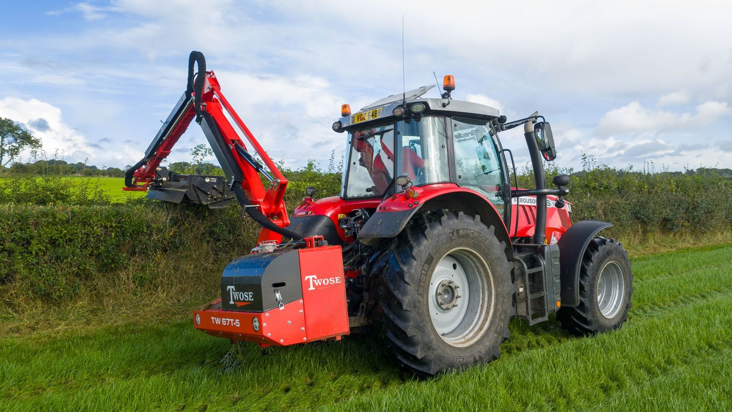 Red agricultural tractor with boom mower attachment working in green field under cloudy sky.