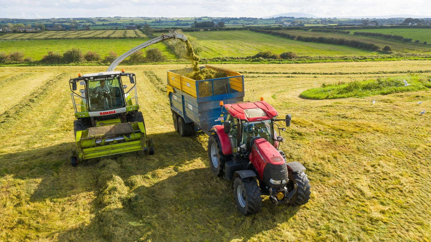A panoramic view of agricultural fields with tractors harvesting crops on rolling green hills during summer.